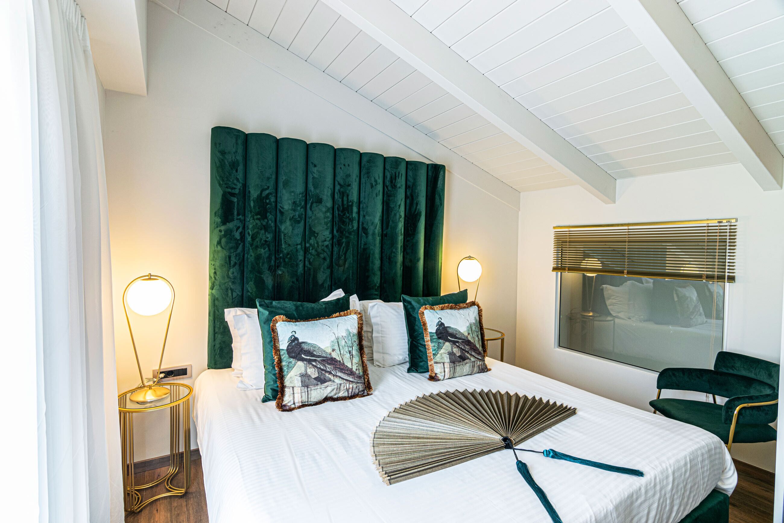 Habitación de hotel con una cama grande, dos lámparas de noche, un cabecero tapizado de color verde y una ventana con persianas.