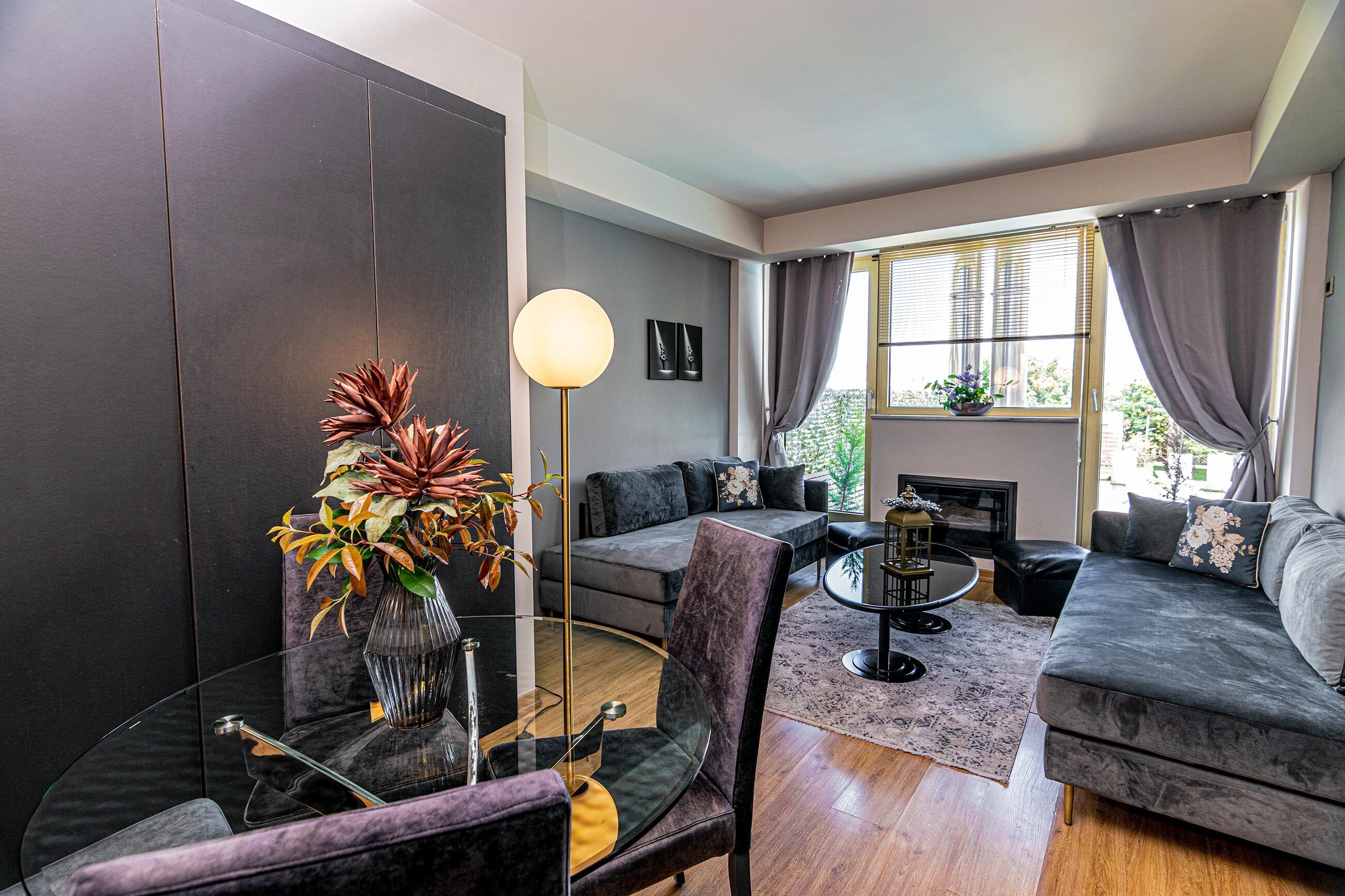 A modern living room with a grey sofa, a glass coffee table, and a dark floor. There is a vase with plants on a side table, and a lamp on a stand.