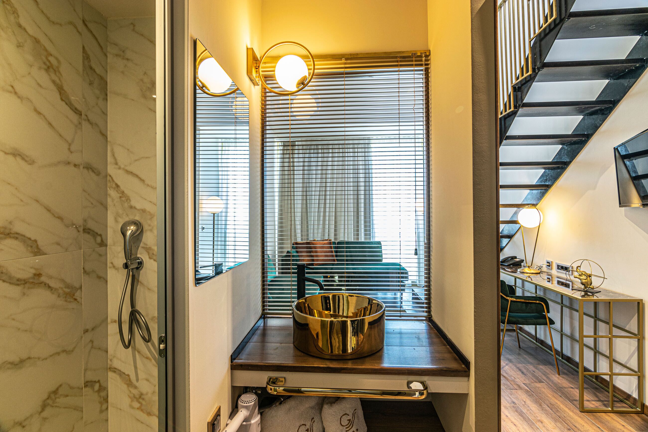 A bathroom with a marble wall, a shower, a sink with a gold basin, and a window with blinds.
