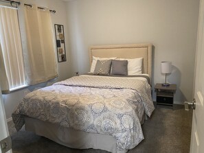 Quarto com cama, mesa de cabeceira, abajur e prateleira na parede.