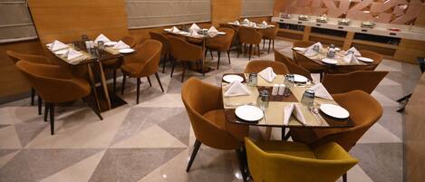 Un comedor con varias mesas y sillas, dispuesto en un ambiente moderno con un patrón geométrico en el suelo y un muro decorativo.
