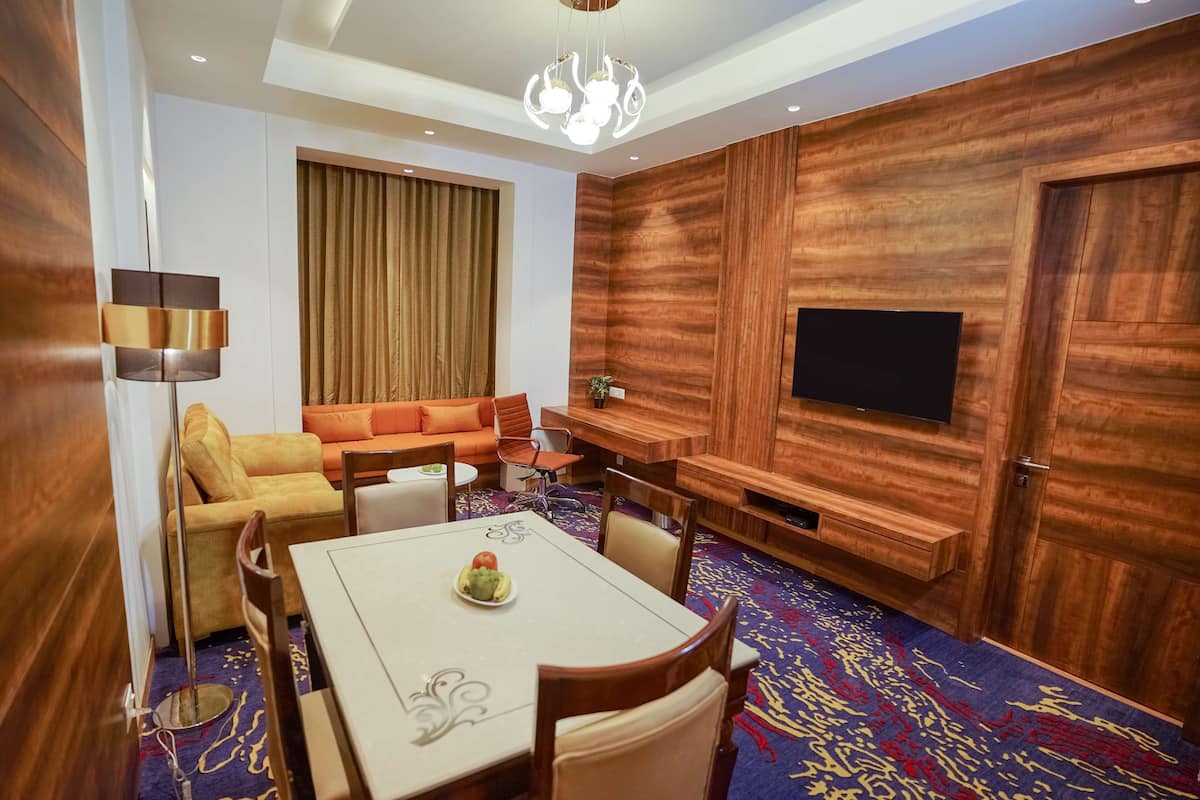 Ein modernes Hotelzimmer mit einer Wandpaneele aus Holz, einem Flachbildfernseher, einem Sofa, einem Esstisch mit Stühlen und einem gemusterten Teppich.