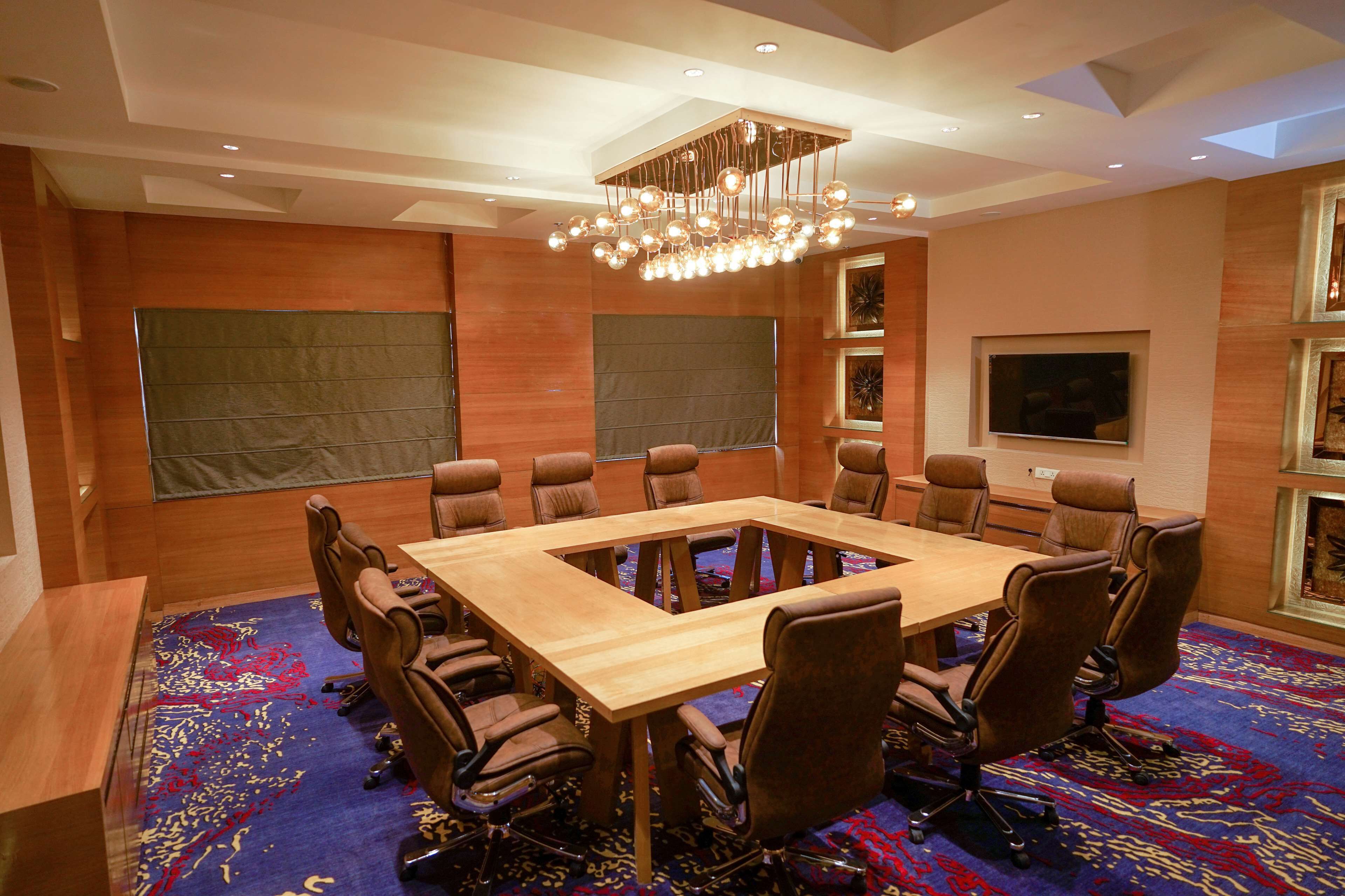 Uma sala de conferências com uma grande mesa de madeira, cadeiras de couro marrom, um candelabro e um tapete estampado.