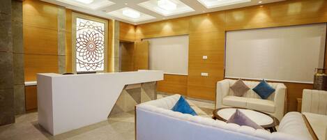 A modern reception area with a white reception desk, a seating area with white sofas and a round table, and a decorative wall panel.