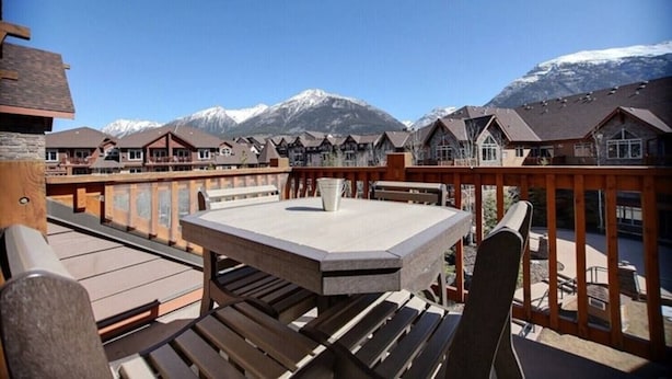 A wooden deck with a table and chairs, overlooking a row of chalet-style buildings with snow-capped mountains in the background.