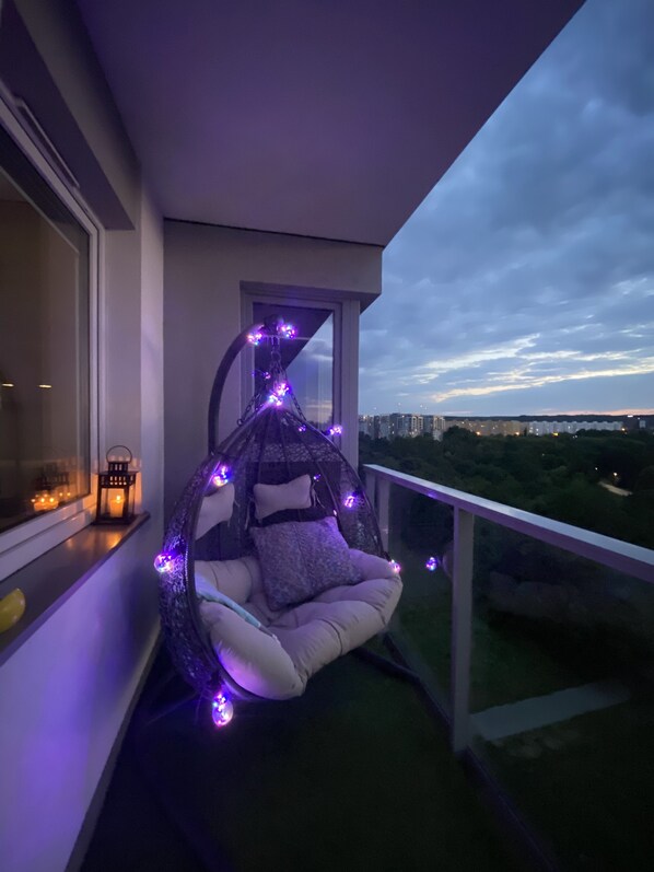A balcony with a hanging chair decorated with purple lights, a lantern, and a view of the cityscape.