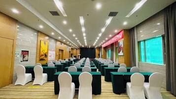 A conference room with rows of tables and chairs, a stage at the front, and a large window on the right.