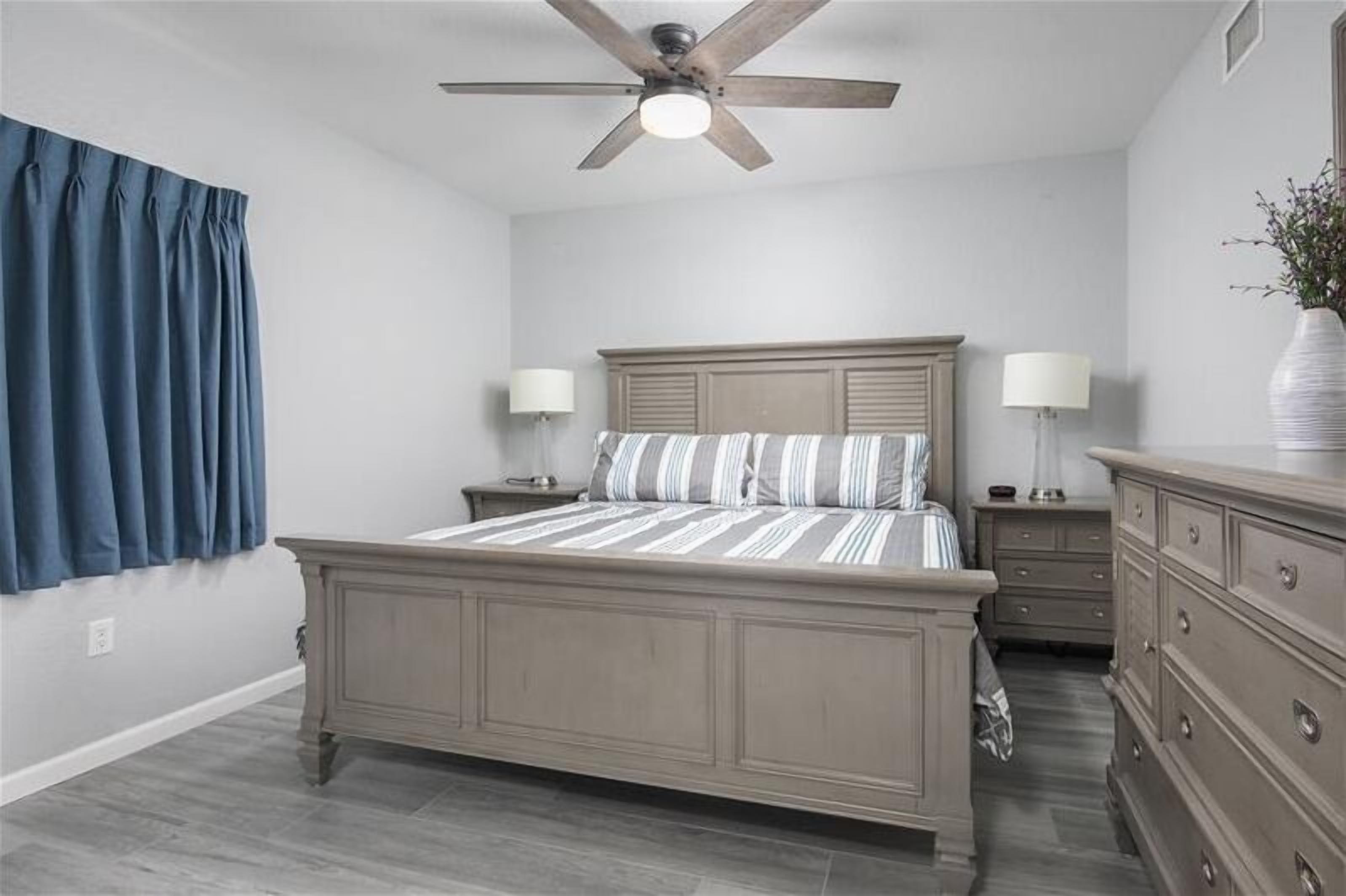 Une chambre à coucher comprenant un lit, des tables de chevet, une commode, un ventilateur de plafond et une fenêtre avec des rideaux.