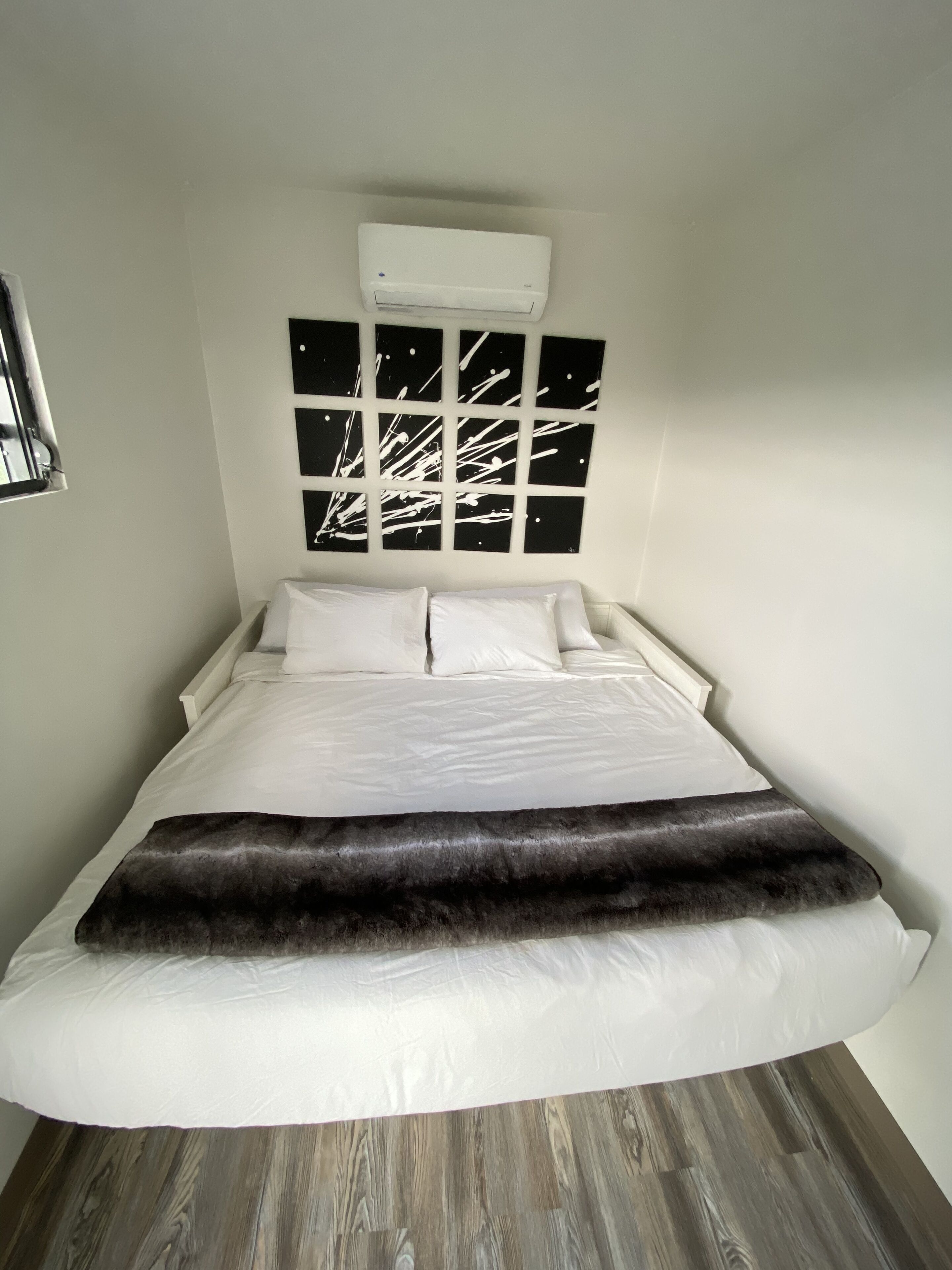 Un dormitorio con cama, ropa de cama blanca, un plaid de piel sintética negro, aire acondicionado de pared y una obra de arte abstracta en blanco y negro sobre la cama.
