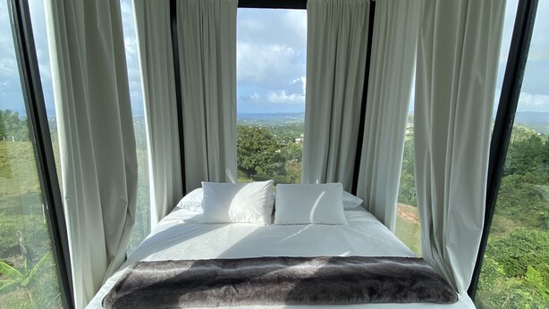 Une chambre à coucher avec un grand lit, du linge de lit blanc et une vue sur un paysage verdoyant grâce à de grandes fenêtres du sol au plafond.