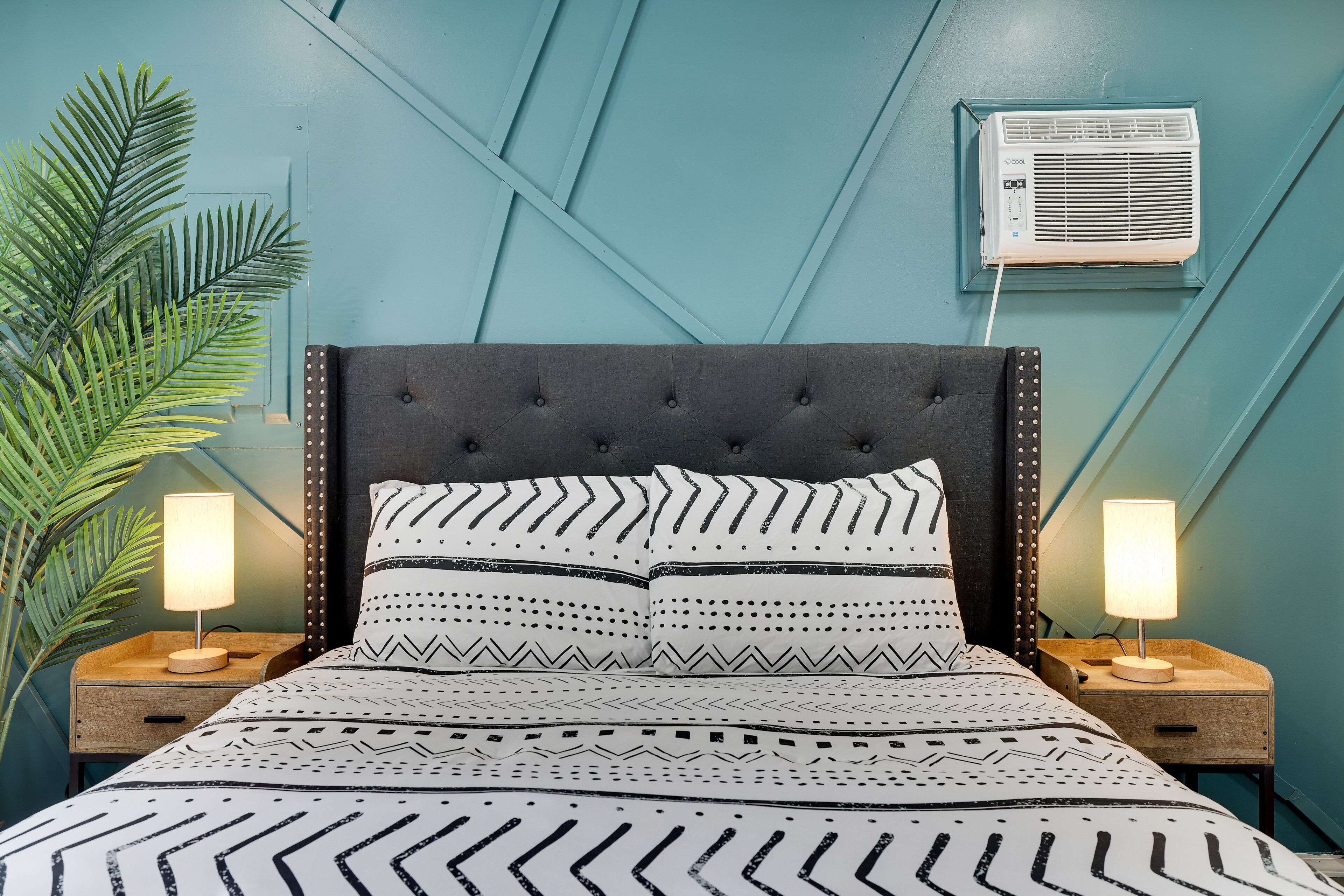 A bed with a patterned bedspread, two bedside lamps, a wall-mounted air conditioner, and a large plant.