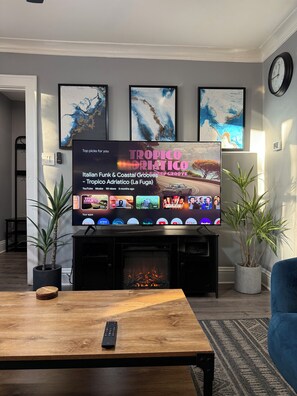 Een woonkamer met een flatscreen televisie waarop een muziekzender wordt vertoond, een open haard, een salontafel en twee potplanten.
