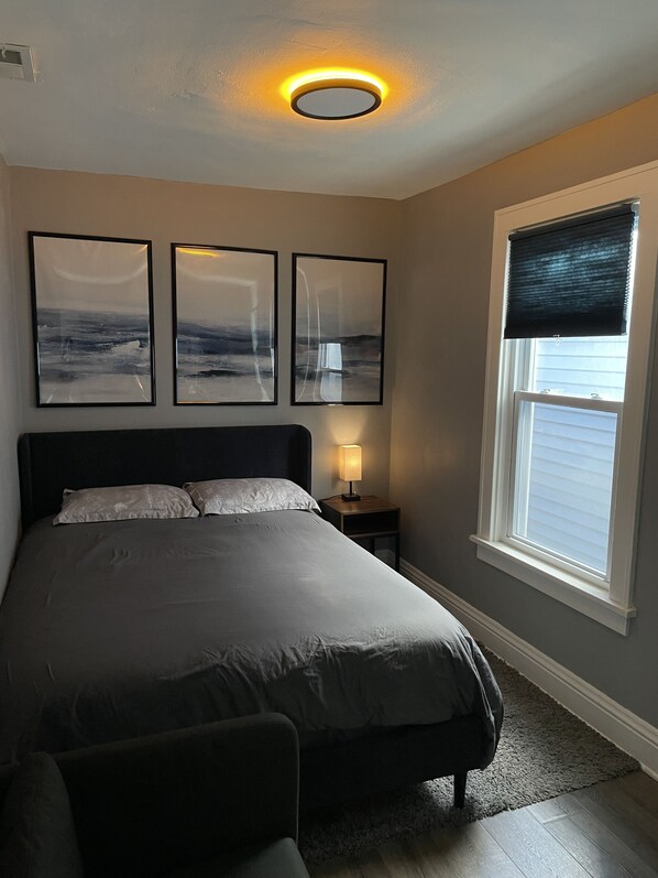 Een slaapkamer met een bed, een nachtkastje met een lamp, drie ingelijste abstracte schilderijen aan de muur, en een raam met jaloezieën.