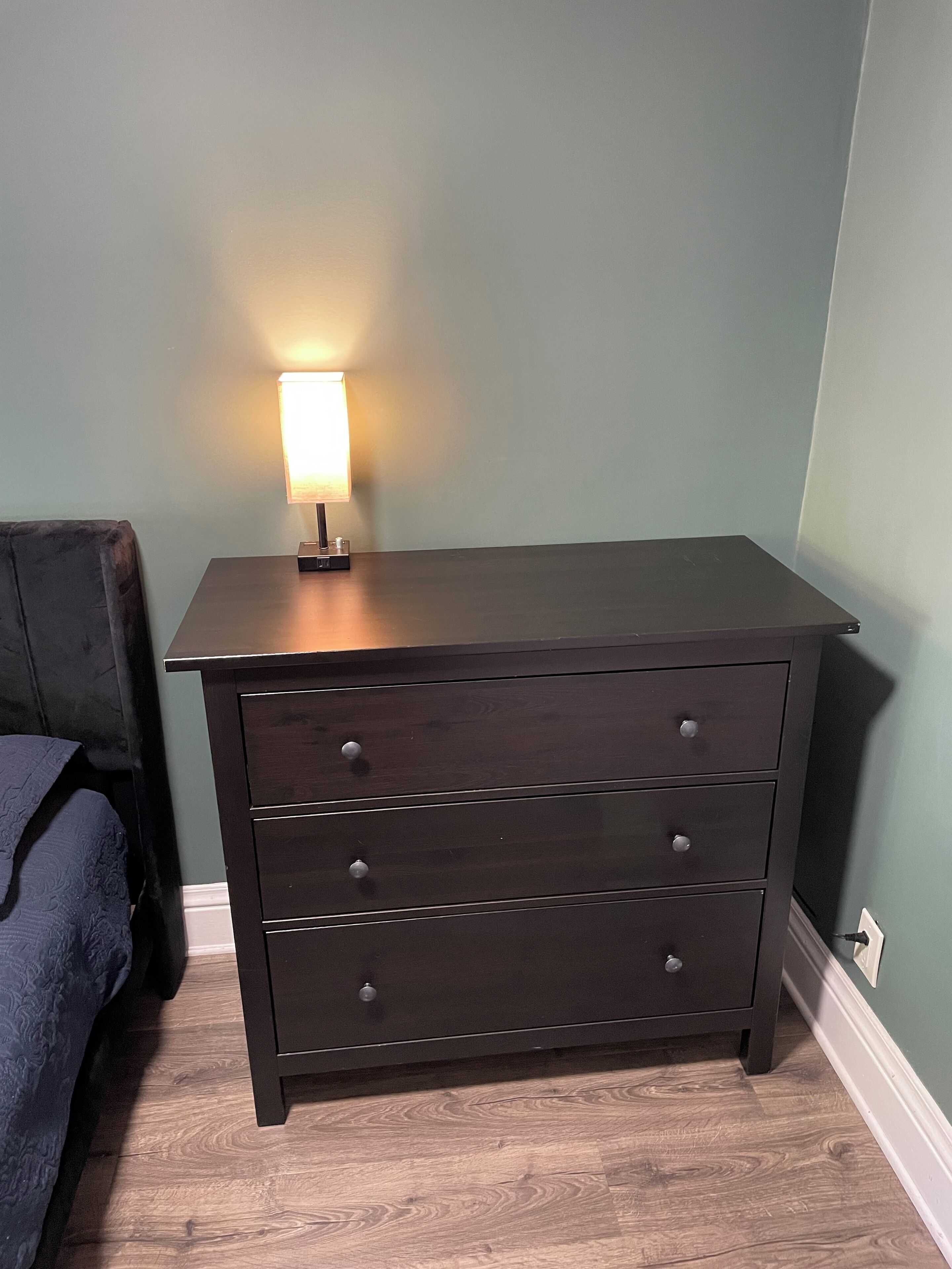 A dark wooden dresser with three drawers and a lamp on top, positioned against a green wall with a wooden floor.