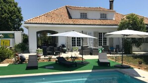 Un área de piscina con sillones de descanso y sombrillas, un patio con mobiliario exterior y una casa con tejado de baldosas.