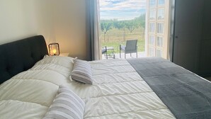 Un dormitorio con cama, mesita de noche, lámpara y puerta abierta que da a un balcón con vistas a un viñedo.