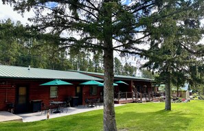 A log cabin with outdoor seating, a covered patio, and a view of surrounding trees. - Whitetail Creek Camping Resort (Lead)