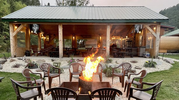 A wooden outdoor seating area with a fire pit surrounded by plastic chairs. - Whitetail Creek Camping Resort (Lead)