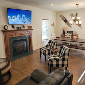 Una sala de estar con chimenea, televisor montado en la pared, comedor con mesa y sillas, y candelabro.