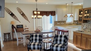 A dining area with a round glass table, two checkered chairs, a chandelier, and a kitchen in the background.