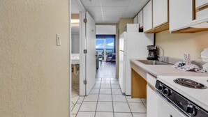 Cocina estrecha con armarios blancos, estufa y cafetera. Se ve una vista del balcón a través de una puerta abierta.