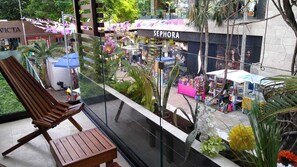 Een balkon met een houten stoel en een tafel, met uitzicht op een straat met winkels, waaronder Sephora.