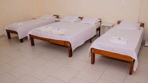 A hotel room with four single beds, each with a white bedspread and pillows, and a wooden headboard. The room has a tiled floor and plain walls.
