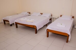 A hotel room with four single beds, each with a white bedspread and pillows, and a wooden headboard. The room has a tiled floor and plain walls.