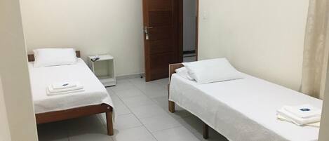 A small, clean room with two single beds, a wooden door, and a white tiled floor.
