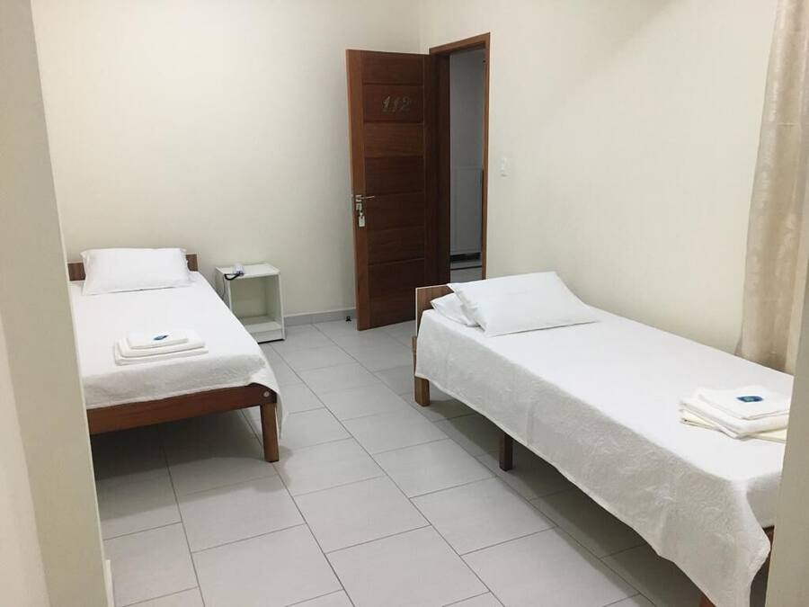 Quarto pequeno e limpo, com duas camas de solteiro, porta de madeira e chão com azulejos brancos.