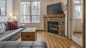 Un salon avec une cheminée en pierre, un canapé gris, une table basse en bois et un téléviseur fixé au-dessus de la cheminée.