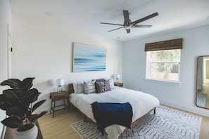 Une chambre à coucher comprenant un lit, des tables de chevet, un ventilateur de plafond, une fenêtre avec des stores et un tableau accroché au mur.