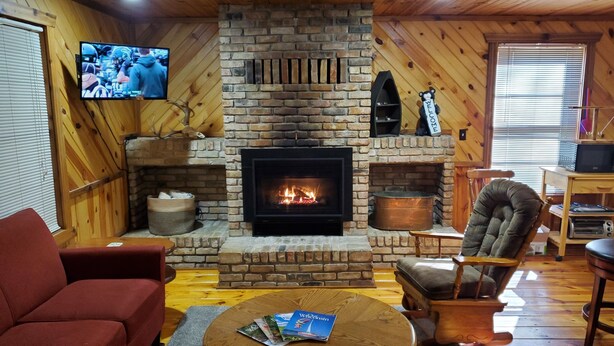 A cozy living room with a brick fireplace, a television mounted on the wall, and a comfortable seating area with a red sofa and a wooden armchair. - Toddy's Cabin (Trempealeau)