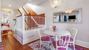 Un área de comedor con una mesa redonda, sillas blancas y un tapete floral. Se ve una escalera con barandillas de madera al fondo.