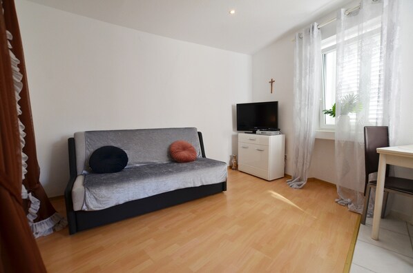 A modern living room with a grey sofa, a flat-screen TV, a small cabinet, and a window with sheer curtains.