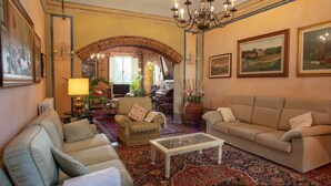A spacious living room with a large sofa set, a coffee table, and a fireplace. There are several framed pictures on the walls and a chandelier hanging from the ceiling.