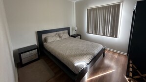 A bedroom with a bed, a nightstand, a ceiling fan, a window with blinds, and a wooden floor.