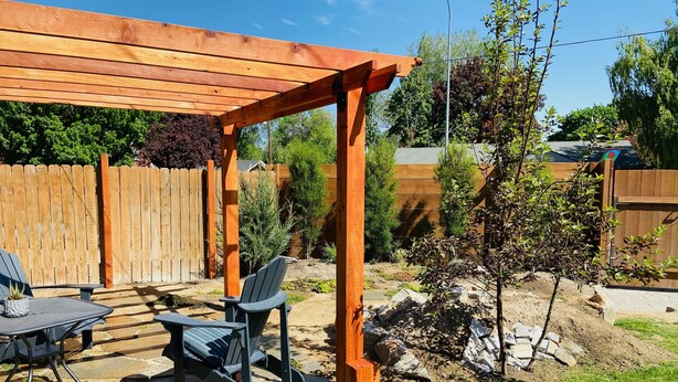 En pergola av trä med ett bord och stolar, omgiven av en trädgård med olika växter och ett litet träd.