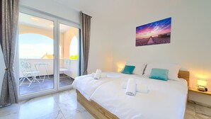 A hotel room with a bed, a painting, a balcony with a table and chairs, and a view of the sky.