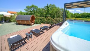 A wooden deck with lounge chairs and a hot tub, surrounded by greenery and a clear sky.
