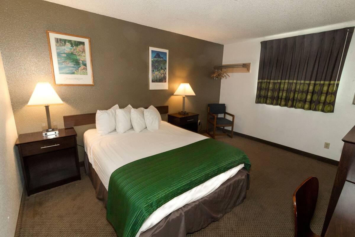 Habitación de hotel con cama, dos mesitas de noche, dos lámparas, una silla y dos cuadros en la pared.