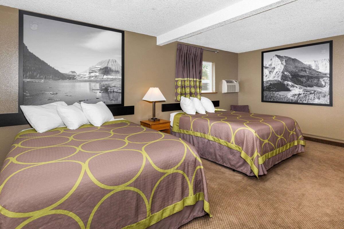 Habitación de hotel con dos camas, una mesita de noche, una lámpara y dos fotografías de paisajes grandes en las paredes.