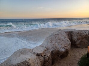 A beach with waves crashing onto the shore, a rocky outcrop in the foreground, and a clear sky.
