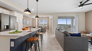 Et moderne køkken med en central ø, spiseområde, samt en stue med sofa og udsigt til det fri.