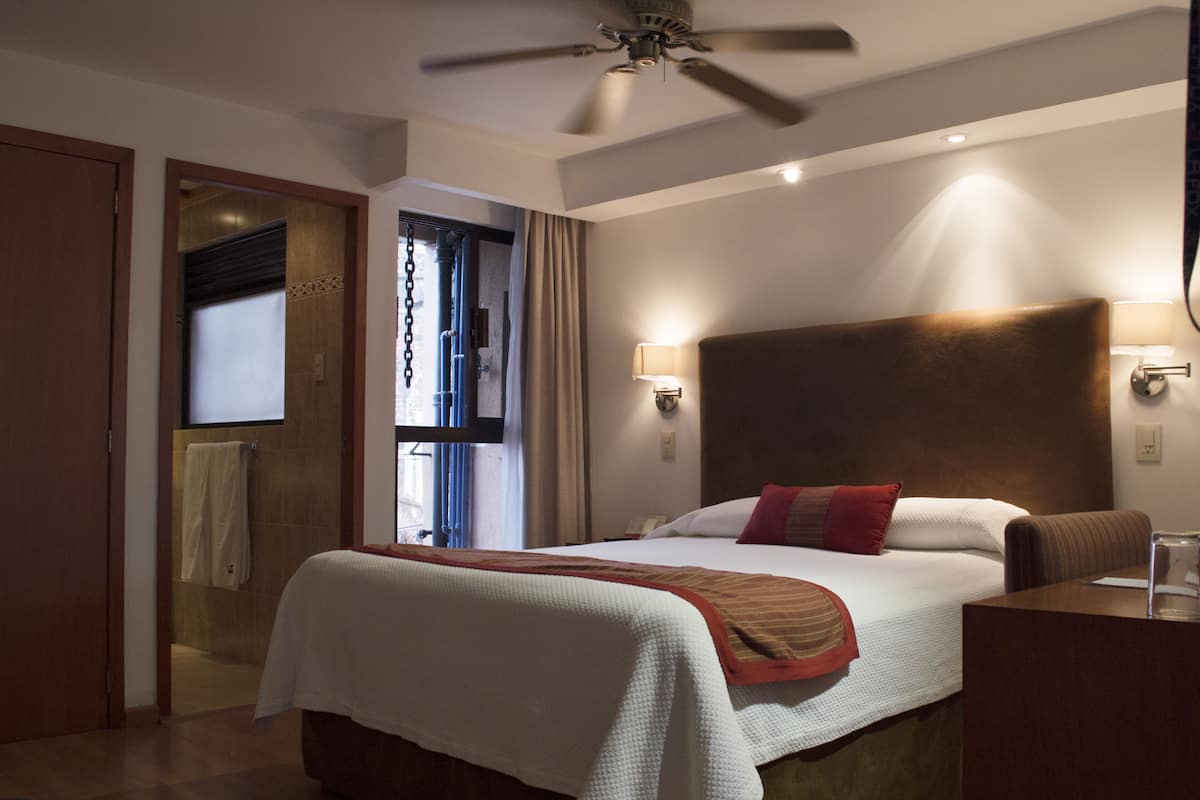 Una habitación de hotel con cama, ventilador de techo, mesita de noche y ventana con cortinas.