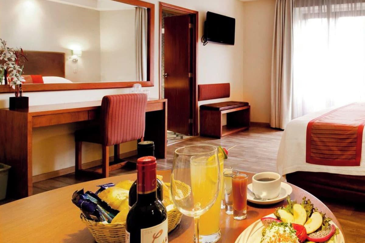 Una habitación de hotel con cama, escritorio y silla, televisión, y una mesa de comedor con comida y bebidas.