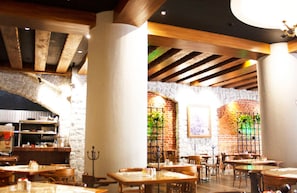 Interior de restaurante con vigas de madera a la vista, paredes de piedra y detalles de ladrillo. Mesas y sillas dispuestas para comer.