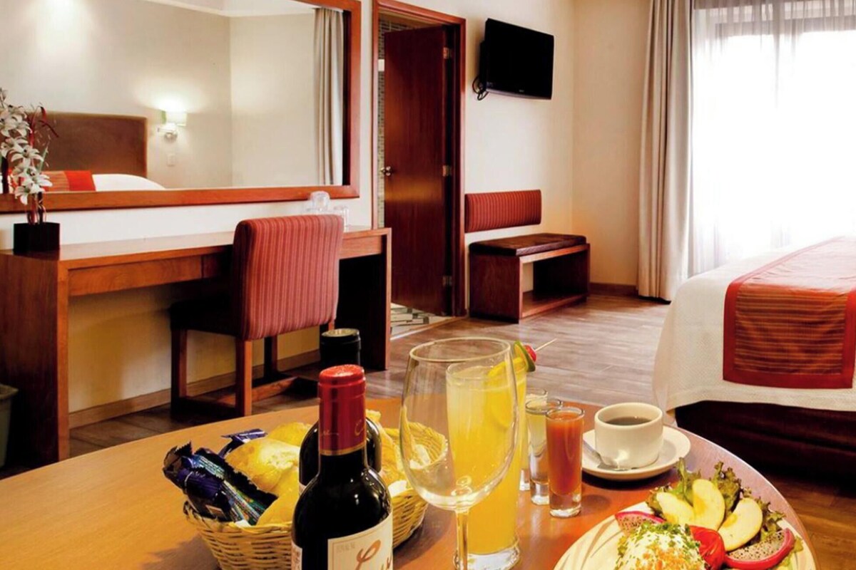 Una habitación de hotel con cama, escritorio, silla, televisión y mesa de desayuno con comida y bebidas.