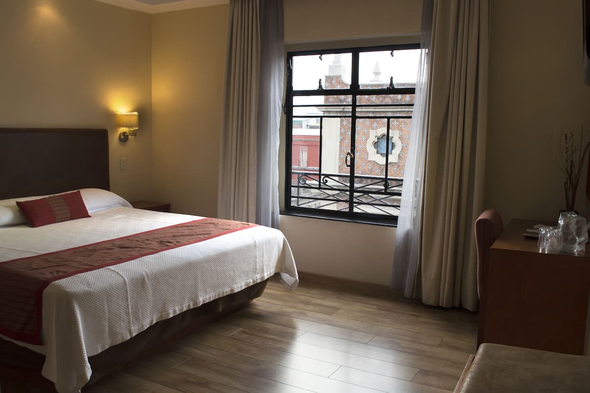 Habitación de hotel con cama, escritorio, silla, ventana con vista a edificios y una lámpara de pared.