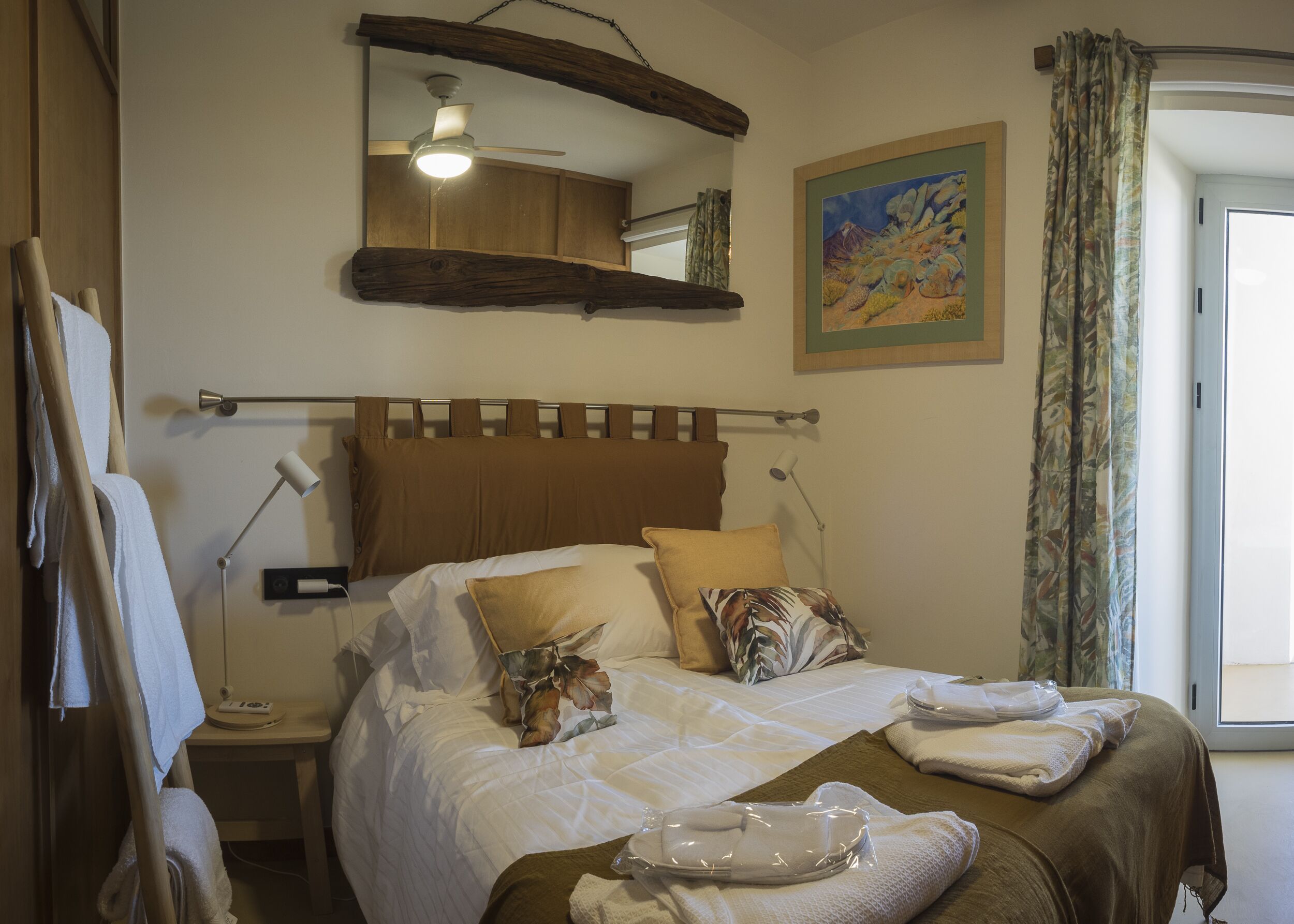 A neatly made bed with a headboard, a bedside lamp, a painting, and a window with curtains.